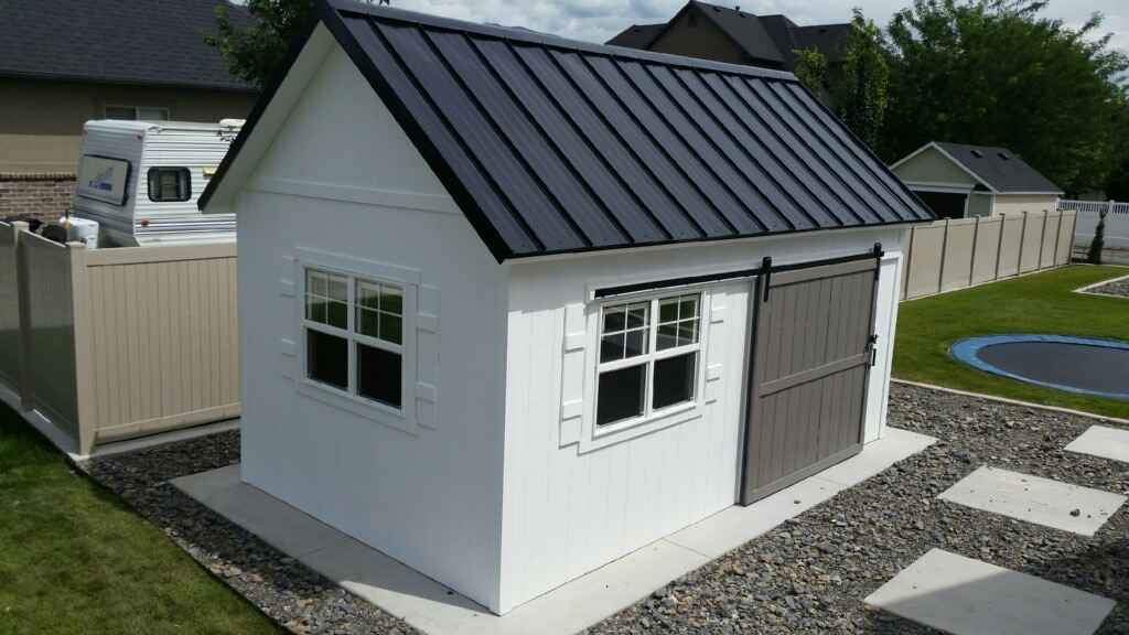 A white orchard shed with a black roof and a stylish sliding barn door, featuring two windows.