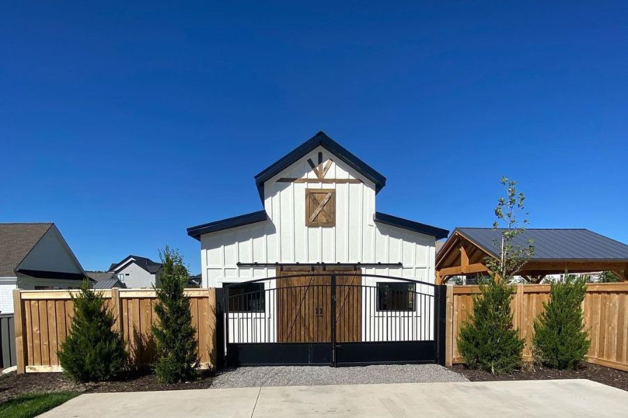 Image of a white and black pole barn with wooden accents, gated entrance, and a blue sky background.
