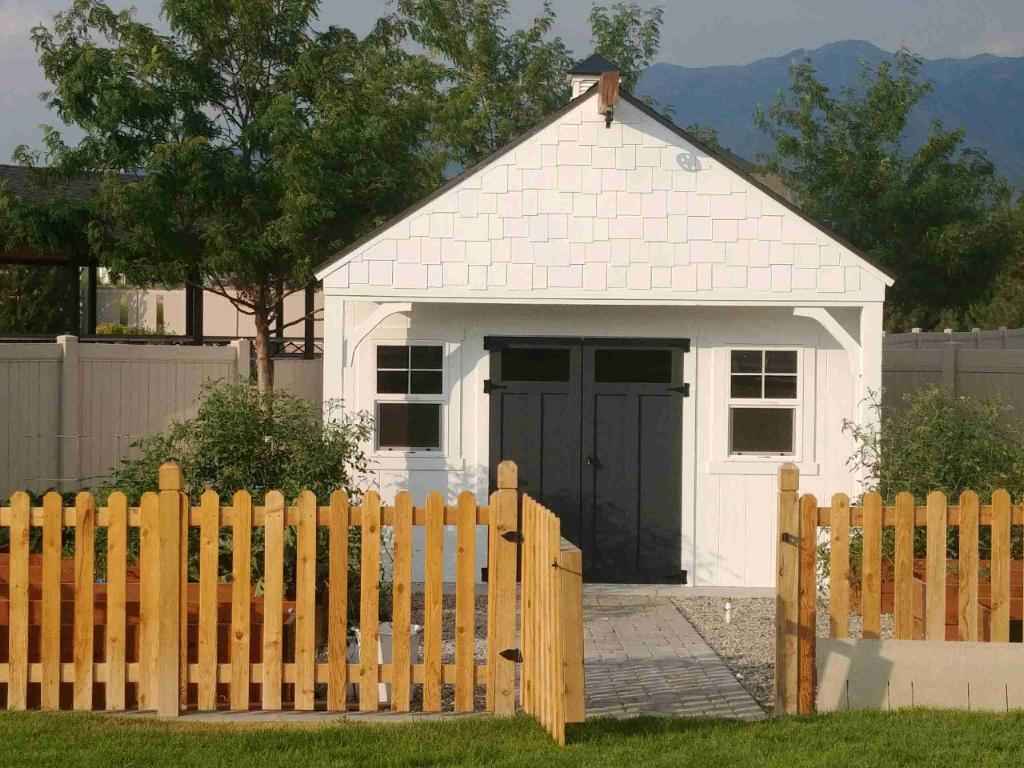A beautiful custom gable shed with a shake shingle roof, white siding, and a black door.
