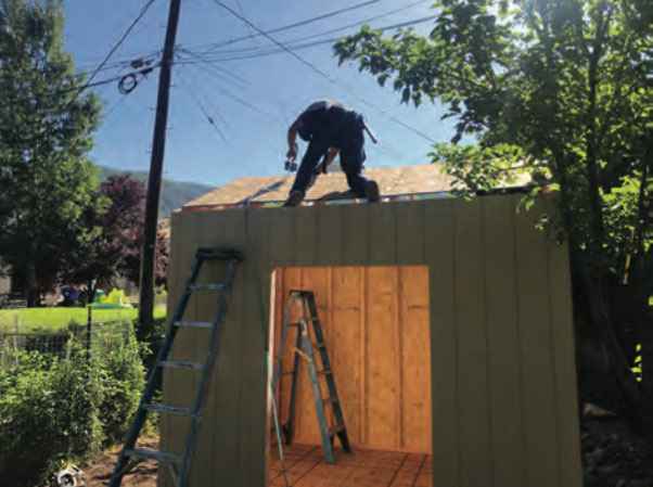 A worker installing the roof of a custom shed while standing on top, with ladders nearby.